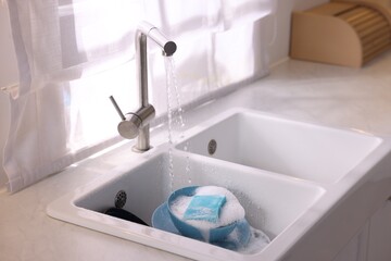 Dishes, blue sponge and foam under tap water in kitchen sink
