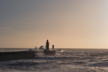 A lighthouse and a concrete pier on the shore of a raging ocean. Huge waves crash against the...