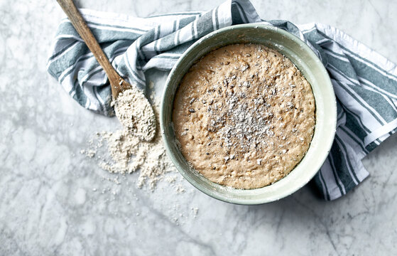 Sourdough bread dough rising in a ceramic bowl on a marble background with flour and wooden spoon