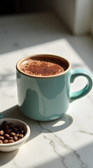 A tight shot of a pastel blue ceramic mug with a thick handle, filled with rich hot cacao topped with foam, sits beside a small bowl of coffee beans on a marble surface