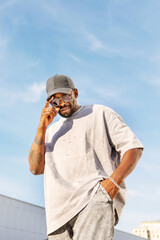 Stylish African American man wearing casual streetwear, sunglasses and Hat while standing outdoors against bright blue sky, looking camera on sunny day. Male fashion model