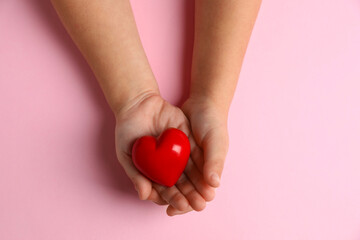 Child holding decorative red heart on light pink background, top view