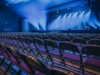 Rows of empty chairs face a stage illuminated by dramatic blue and white lighting. The setup suggests anticipation for an upcoming event or performance in a concert hall.
