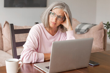 Sad upset, gray-haired, middle-aged woman is sitting in front of a laptop