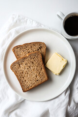 A photograph of an elegant, minimalist arrangement featuring two slices of whole-grain bread with butter and coffee on the side