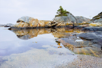 Beautiful rocks and pine tree reflected in calm coastal waters, stones mirroring in still water. Serene Scandinavian atmosphere. For meditation visuals, travel blogs and nature backgrounds