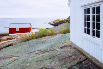 White wooden lighthouse wall over the sea and classic red boathouse nearby. Idyllic Nordic coastal landscape. For maritime heritage features, travel inspiration and coastal conservation themes. © Nimbus Works
