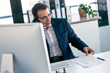 Cheerful businessman talking on mobile phone while working with computer in the office