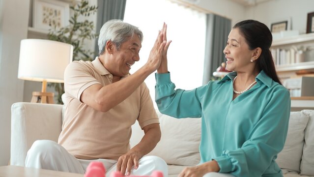Elder woman using dumbbell at living room for boosting balance and coordination or enhancing joint health and flexibility while grandfather encouraging grandmother and giving high five. Myrmidon.