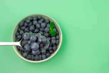 A spoonful of blueberries in a bowl. The bowl is white and the spoon is silver. The blueberries are fresh and ripe. Green background. Table top view