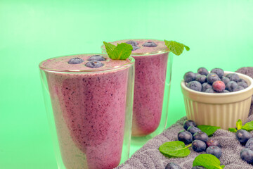 Two glasses of blueberry smoothies with mint leaves on top. A bowl of blueberries on the side. Green background