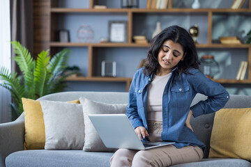 Woman experiencing back pain while sitting with laptop on the couch at her home office.