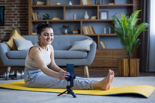 Young woman records a workout session on her phone, sitting on a yellow mat in her living room. - Powered by Adobe