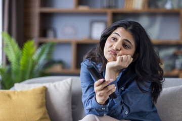A bored young woman rests her head on her hand while watching tv with the remote control.