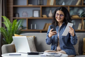 Young woman happily manages finances at home using her smartphone for convenient bill payments.