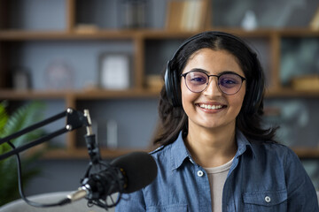 A young woman with headphones smiles in front of a microphone, ready to record a podcast.
