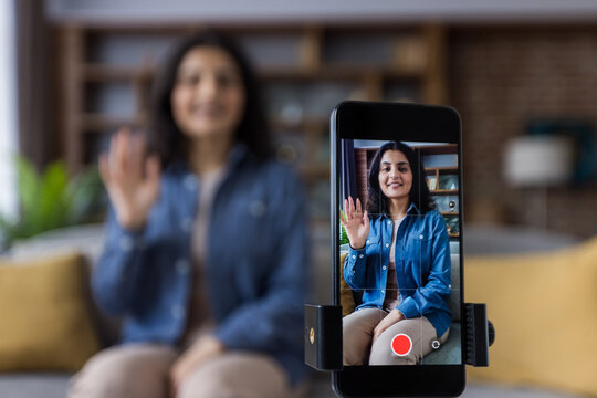 A smiling woman waves to the camera while recording a video with her smartphone on a tripod.