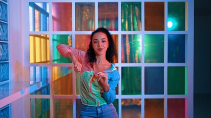 Happy hispanic girl in lively mood moving along music in neon light while wearing colorful cloth at night club. Street dancer making energetic footstep or movement while looking at camera. Regalement.