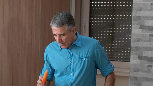 Portrait of a Handsome 54-year-old Man with Gray Hair in Shirt Preparing Food at Home. Cheerful man uses a vegetable slicer to slice carrots. Perfect for promoting lifestyle of middle-aged men.