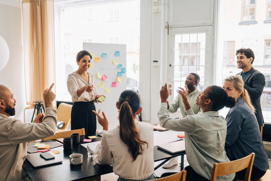 Smiling female entrepreneur discussing and asking business plans to colleagues sitting at table in office meeting room