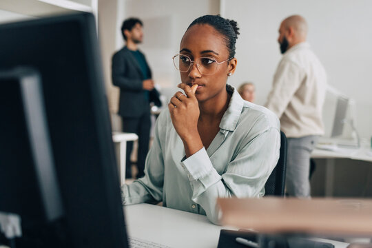 Contemplative young creative professional wearing eyeglasses sitting at desk while working on desktop PC in office