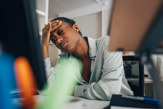 Tired young businesswoman wearing eyeglasses sitting at desk with eyes closed in office