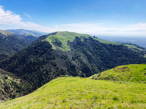 A scenic view of Alum Rock Park rugged terrain highlights vibrant grass covered slopes with hiking trails, shadowed valleys, and distant urban San Jose sprawl framed by foothills of the Diablo Range