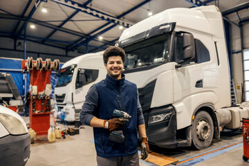 Fototapeta premium Portrait of diverse mechanic with drill in hands standing at mechanic workshop and preparing for lorry fixing.