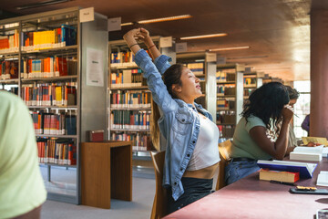 Female student stretching with arms raised while sitting at desk in university library