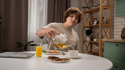 Young man in the morning in the kitchen dinning room at home enjoying healthy breakfast, sitting at table pouring herbal tea in the cup.
