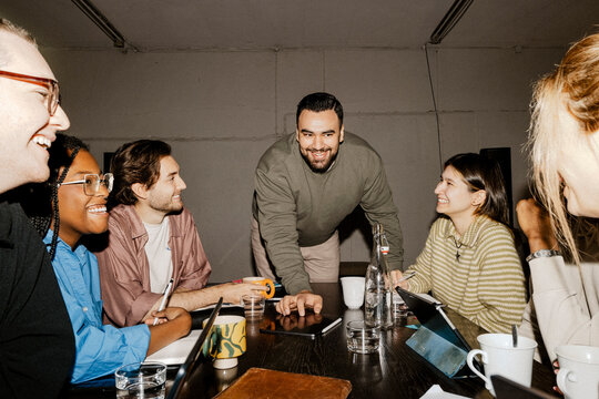 Smiling male entrepreneur leaning on table and talking with team in meeting at office