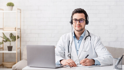 Online communication with patients. Serious doctor in white coat and headphones sits at table with...