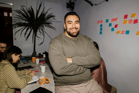 Portrait of smiling male entrepreneur sitting on table with arms crossed at office
