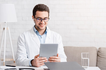 Online visit to doctor. Man in glasses and white coat looks at tablet, sitting at table with...