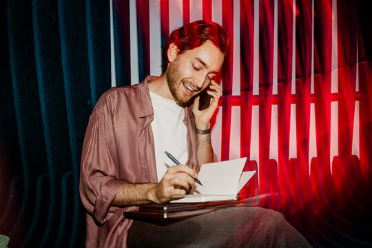 Smiling male entrepreneur talking on smart phone and writing in diary while sitting on seat