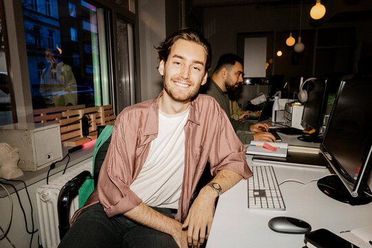 Smiling young male entrepreneur sitting at desk in tech start-up office