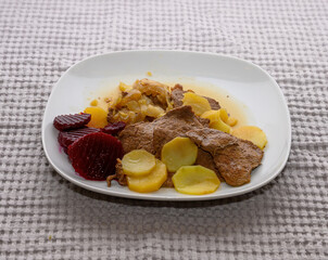 Savory beef slices are accompanied by potatoes and beetrot arranged neatly on a white plate. The hearty meal is set against a soft, textured tablecloth, inviting comfort
