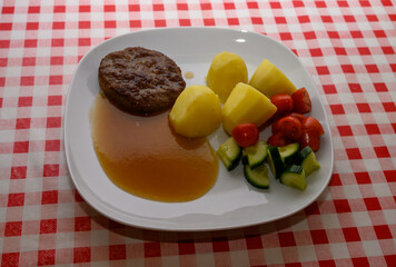 A hearty meal is presented with a meat patty beside boiled potatoes, drizzled with savory sauce, and accompanied by fresh tomatoes and cucumbers on a checkered tablecloth