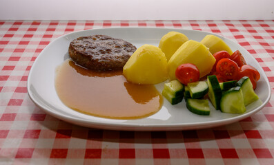 A hearty meal is presented with a meat patty beside boiled potatoes, drizzled with savory sauce, and accompanied by fresh tomatoes and cucumbers on a checkered tablecloth