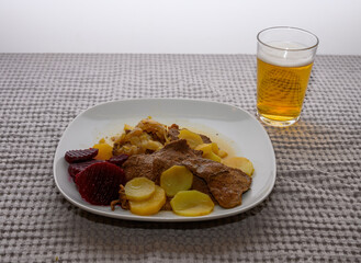 Savory beef slices are accompanied by potatoes and beetrot arranged neatly on a white plate. The hearty meal is set against a soft, textured tablecloth, inviting comfort