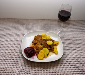 Savory beef slices are accompanied by potatoes and beetrot arranged neatly on a white plate. The hearty meal is set against a soft, textured tablecloth, inviting comfort