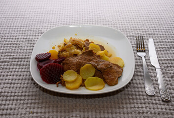 Savory beef slices are accompanied by potatoes and beetrot arranged neatly on a white plate. The hearty meal is set against a soft, textured tablecloth, inviting comfort