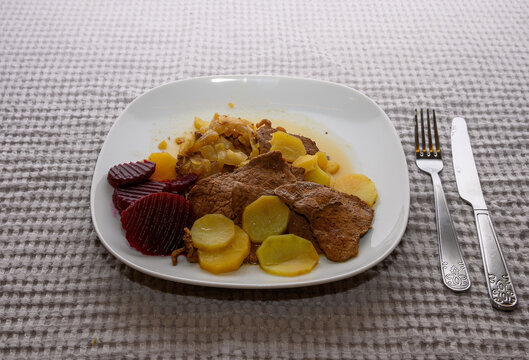 Savory beef slices are accompanied by potatoes and beetrot arranged neatly on a white plate. The hearty meal is set against a soft, textured tablecloth, inviting comfort