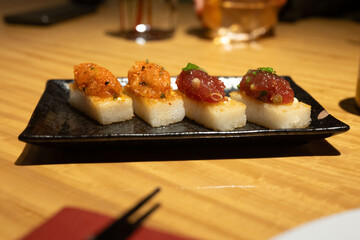 Artisan sushi bites on a sleek black plate, featuring rice cakes topped with fresh seafood and flavorful garnishes, set against a warm background.