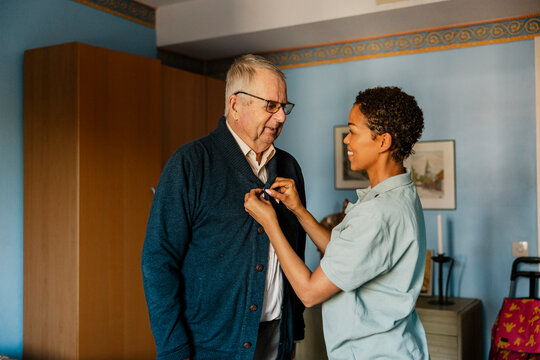 Smiling female caregiver buttoning jacket of senior man at home
