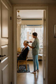 Side view of female nurse combing hair of senior woman at home seen through doorway