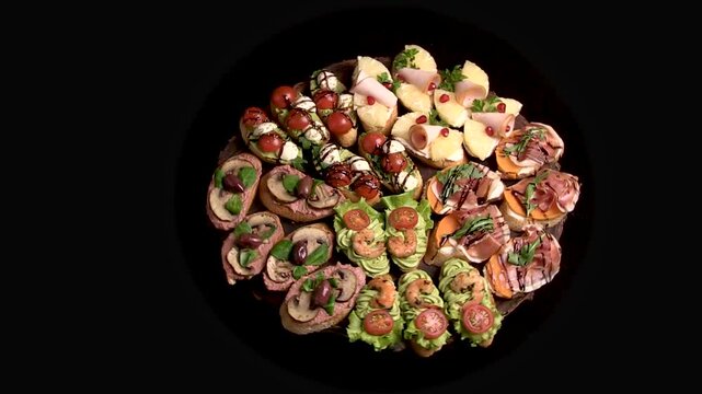 An appetizer board with various bruschetta for a buffet: jamon, ham, pate, tomatoes, cheese. The appetizer plate rotates on a black background. Top view, general view