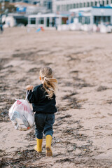 Little girl with a bag of garbage walks along the beach towards apartment buildings. Back view