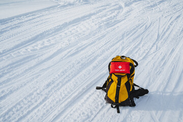 A bright yellow backpack sits prominently in the snow, accompanied by a striking red first aid kit, highlighting the adventure and preparations needed for winter activities