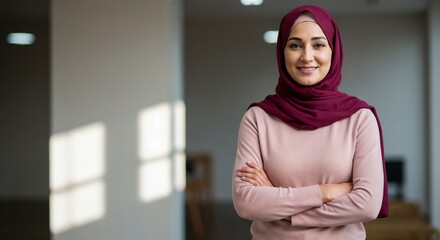 A young woman in a maroon hijab stands confidently indoors, smiling at the camera with her arms crossed. Natural light from the windows highlights her modest yet stylish attire